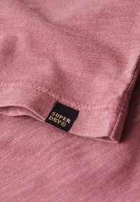 Rosa bomullstyg med en subtil textur, med en mörk etikett som har texten "SUPER DRY" i guld. Enkelt mönster, slät yta.