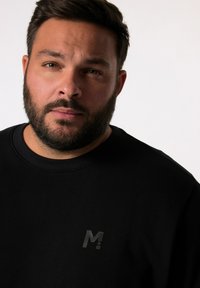 Sudadera negra con cuello redondo, con un pequeño logo "M" negro brillante en el pecho. La tela parece suave y lisa.