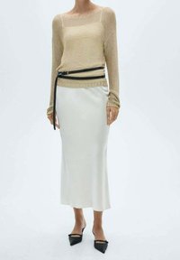 Femme portant un pull beige en maille, ceinturé à la taille, associé à une jupe midi blanche et des ballerines noires à bout pointu.