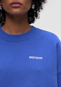 Niebieska bluza z okrągłym dekoltem, ściągaczami przy rękawach oraz haftowanym logo "WRSTBHVR" na klatce piersiowej, wykonana z miękkiego, strukturalnego materiału.