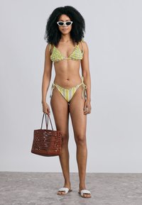 Bikiny so zeleným a žltým vrchným dielom s volánikmi, pruhované spodné diely; doplnky zahŕňajú biele slnečné okuliare a hnedú pletenú tašku.
