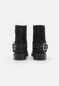 Bottines en daim noir avec un talon plat, dotées d'une sangle en cuir et d'un détail de boucle en métal. Texture lisse avec des surpiqûres contrastées.
