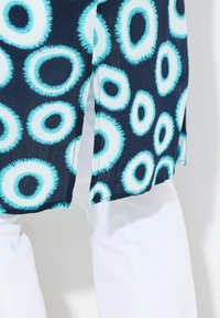 Modrý vzorovaný textil s kulatými motivy v různých odstínech na námořnickém pozadí, doplněný bílými kalhotami, který ukazuje texturu a design.