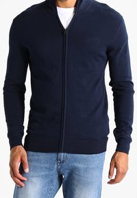 Marinblå zip-up cardigan tillverkad av mjukt sträckt tyg. Har hög krage, ribbade ärmslut och nederkant, samt en minimal logotyp på bröstet.