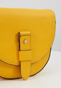 Borsa a tracolla in pelle gialla con patta arrotondata, chiusa da una cinghia con bottone automatico. Presenta cuciture a contrasto e una texture liscia.