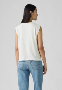Biała bawełniana bluzka bez rękawów o luźnym kroju, pokazana z jasnoniebieskimi dżinsami. Prosty design z okrągłym dekoltem, bez widocznych wzorów.
