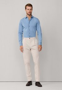 Muškarac u svijetloplavoj košulji s dugim rukavima, bež hlačama i tamnosmešnim cipelama stoji naslonjen uz jednoličnu pozadinu.