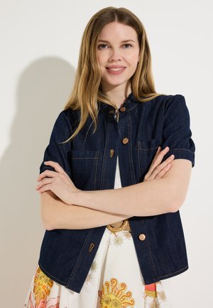 Junge Frau mit langen blonden Haaren, lächelnd, trägt eine kurzärmelige dunkle Jeansjacke über einem weißen Kleid mit bunten Sonnen- und Blumenmustern.