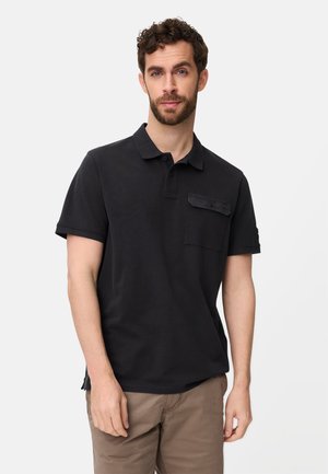 Man met kort krullend haar en baard, gekleed in een zwart poloshirt met korte mouwen en een borstzak, en een bruine broek, staand tegen een witte achtergrond.