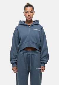 Hoodie grigio corto con chiusura a zip e grande cappuccio, con il testo "GOODFORNOTHING" sul petto, abbinato a pantaloni della tuta con cordino coordinati.