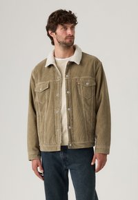 Veste en velours côtelé marron avec un col en polaire blanc, deux poches poitrine et des boutons en argent, associée à un denim noir, sur un fond clair.