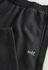 Zwarte sweatpants met een elastische tailleband, twee zijzakken en een klein geborduurd "self." logo in het wit. Glad, zacht materiaal.
