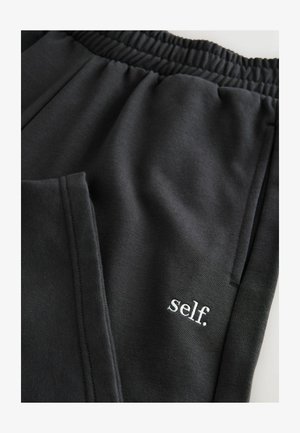 Black sweatpants featuring an elastic waistband, two side pockets, and a small embroidered "self." logo in white. Smooth, soft fabric.