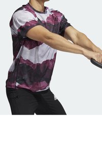 Kortärmad träningsskjorta i ett lila och vitt abstrakt mönster, tillverkad av andningsbart tyg; ihop med svarta träningsbyxor.