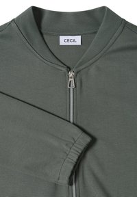 Veste verte à manches longues avec fermeture éclair sur le devant, poignets élastiques et une étiquette blanche marquée "Cecil" à l'intérieur du col.