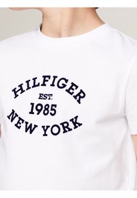 Witte katoenen T-shirt met een marineblauw circulair logo met de tekst "HILFIGER EST. 1985 NEW YORK". Korte mouwen en een ronde halsontwerp.