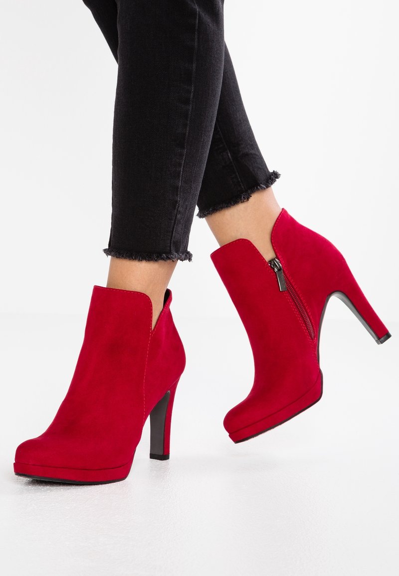 Rote Wildleder-Ankle Boots mit einem hohen Absatz, seitlichem Reißverschluss und Cut-out-Design. Kombiniert mit schwarzen Jeans mit fransigen Säumen.