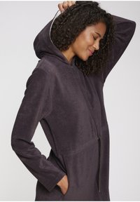 Kapuča, zadrgo zaprt kombinezon v temno sivi barvi, izdelan iz mehke, teksturirane tkanine. Ima dolge rokave in stranske žepe za dodatno praktičnost.