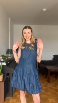 Γυναίκα με μπλε φόρεμα που χαμογελά και κουνά τα χέρια της σε ένα σαλόνι με φυτά, πιάνο, φωτιστικό και έναν σκούρο καναπέ στο φόντο.