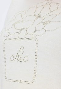 Beige T-Shirt mit floralem Design und dem Wort "chic", das aus kleinen, glänzenden Goldnieten besteht. Weicher Stoff und lässige Passform.