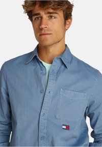 Camicia button-up azzurro chiaro in cotone, caratterizzata da una tasca con il logo Tommy Jeans, una trama sottile e un design del colletto classico.