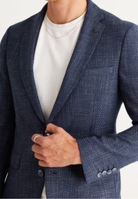 Muž zapíná tmavě modrý texturovaný blazer přes bílou tričko s kulatým výstřihem, nosí tenký zlatý řetízek a stříbrný prsten na levé ruce.