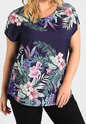 Femme portant un chemisier à manches courtes bleu marine à fleurs avec des fleurs tropicales roses, violettes et vertes et un pantalon noir.