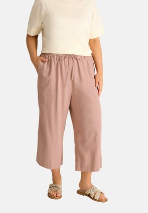 Kobieta ubrana w beżową bluzkę z krótkim rękawem, luźne, jasnofioletowe spodnie trzy czwarte ze sznurkiem oraz plecione beżowe sandały wsuwane, ręce częściowo w kieszeniach.