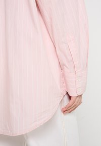 Jasnoróżowa koszula z długim rękawem w cienkie białe pionowe paski, z zaokrąglonym dołem i mankietami zapinanymi na guziki. Miękka, gładka tekstura tkaniny.