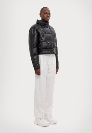 Giacca a vento nera cropped con texture trapuntata, tasche con zip e colletto alto, abbinata a pantaloni bianchi con righe e sneakers bianche.