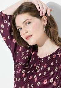 Frau mit heller Haut und blauen Augen, die silberne Creolen trägt und ein lila, floral gemustertes, plissiertes Oberteil hat, während sie sich mit einer Hand das Haar zurückhält.