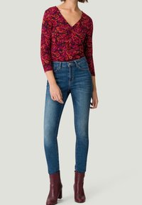 Femme portant un haut à motifs paisley rouge à manches trois-quarts, un jean skinny taille haute bleu et des bottines bordeaux, debout.