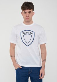 T-shirt bianca in cotone con logo a scudo blu navy e la scritta "Blauer." Maniche corte e scollatura girocollo. Indossata con jeans in denim blu.