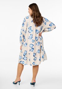 Robe à motifs floraux en beige avec des fleurs aquarelle bleues, manches longues et ourlet froncé. Fabriquée en tissu doux, avec une coupe décontractée.