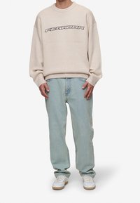 Beige stickad sweatshirt med lös passform och marinblå text "PEGRADOOR." Kombinerad med ljusblå jeans och vita sneakers med beige detaljer.