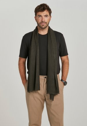 Mann mit kurzen lockigen Haaren, der ein schwarzes T-Shirt, beige Hose, schwarze Uhr und schwarz karierten Schal trägt, steht vor einem schlichten Hintergrund.