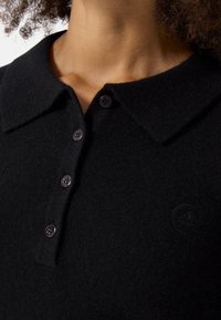 Černé polo tričko vyrobené z jemné tkaniny, s knoflíkovou légou se čtyřmi ladícími knoflíky a vyšívaným logem na hrudi.