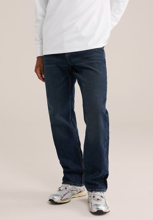 Personne portant un jean bleu foncé, des baskets argentées et blanches, et une chemise blanche à manches longues, debout sur un fond clair.