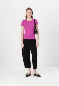 Přiléhavé růžové tričko s potiskem loga, spárované s černými zkrácenými kalhotami a černými slip-on botami. Mini taška visí na popruhu.