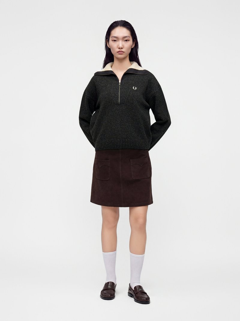 Femme debout, les mains dans le dos, portant un pull zippé foncé, une jupe marron avec poches, des chaussettes blanches et des mocassins foncés sur un fond uni.