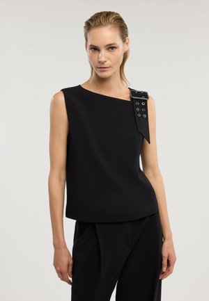 Mujer con el cabello peinado hacia atrás que lleva una blusa negra sin mangas con un escote asimétrico y un gran detalle de hebilla en un hombro.