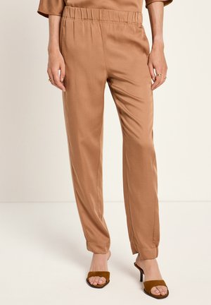 Eine Frau trägt hochgeschnittene, camel-farbene Satin-Hosen mit elastischem Bund und braunen Sandalen mit offenem Zehenbereich und Absatz. Sie steht vor einem einfarbigen Hintergrund.