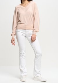 Hellrosa Strickpullover mit V-Ausschnitt, langen Ärmeln und gerafften Bündchen, kombiniert mit weißen, weit geschnittenen Jeans und weißen Sneakers.