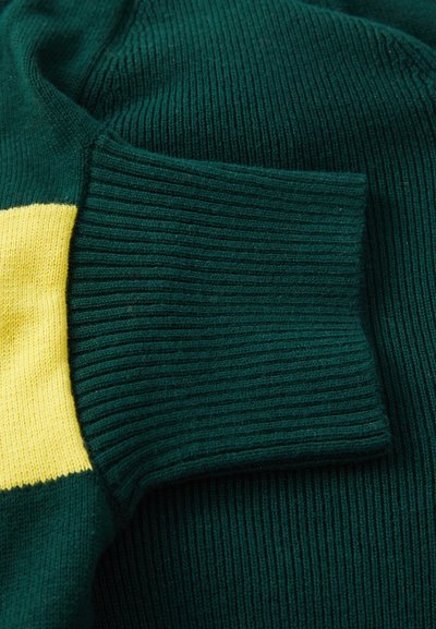 Zeleno pleteno pulover s ribasto teksturo in živahno rumeno vodoravno črto. Podrobnost o manšetah prikazuje kontrastne teksture.