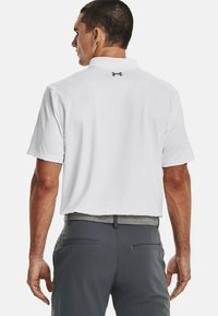 Hvid kortærmet polo shirt med krave, med et sort logo på ryggen. Sammen med grå bukser, der viser lommer og en struktureret talje.