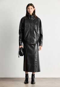 Crna kožna jakna s visokim ovratnikom i narančastim šavovima, u paru s odgovarajućom crnom kožnom suknjom i crnim čizmama s petom.