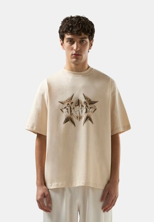 Giovane uomo con capelli ricci che indossa una t-shirt beige oversize con un disegno grafico a forma di stella metallica sul petto.