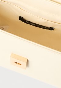 Intérieur d'un sac à main couleur crème montrant une étiquette noire "Anna Field" et une fermeture rectangulaire dorée sur le rabat extérieur.