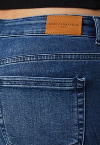 Jeans de mezclilla azul con una textura suave, que presentan un parche de marca de cuero marrón y bolsillos traseros cosidos.