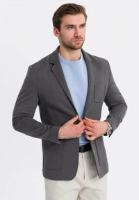 Hombre con un blazer gris sobre un suéter azul claro y pantalones beiges, abrochándose el blazer, frente a un fondo blanco liso.
